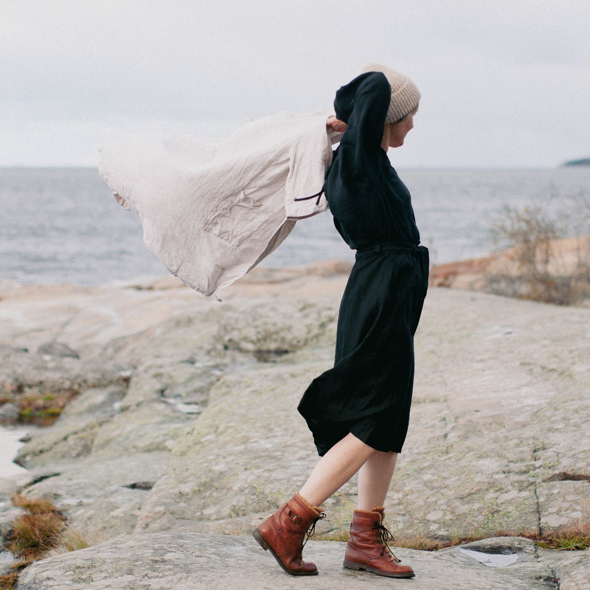 RAORI - En kvinna står vid havet och har en svart kjol och tröja ooch tar på sig en jacka som blåser i vinden.